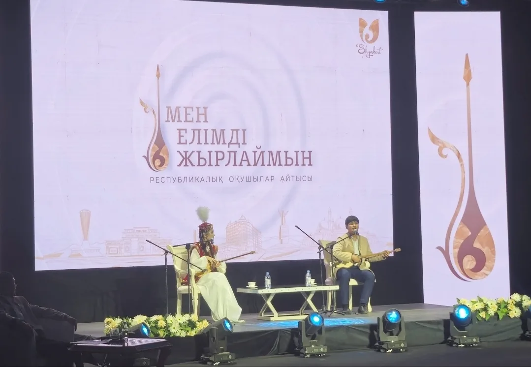 Республиканский айтыс школьников «Мен елімді жырлаймын» в Шымкенте, 6 декабря 2025 года: два юных акына с домбрами на сцене. Фото: TheNomadEditor, Wikimedia Commons, CC0. Временная обложка до своего кадра