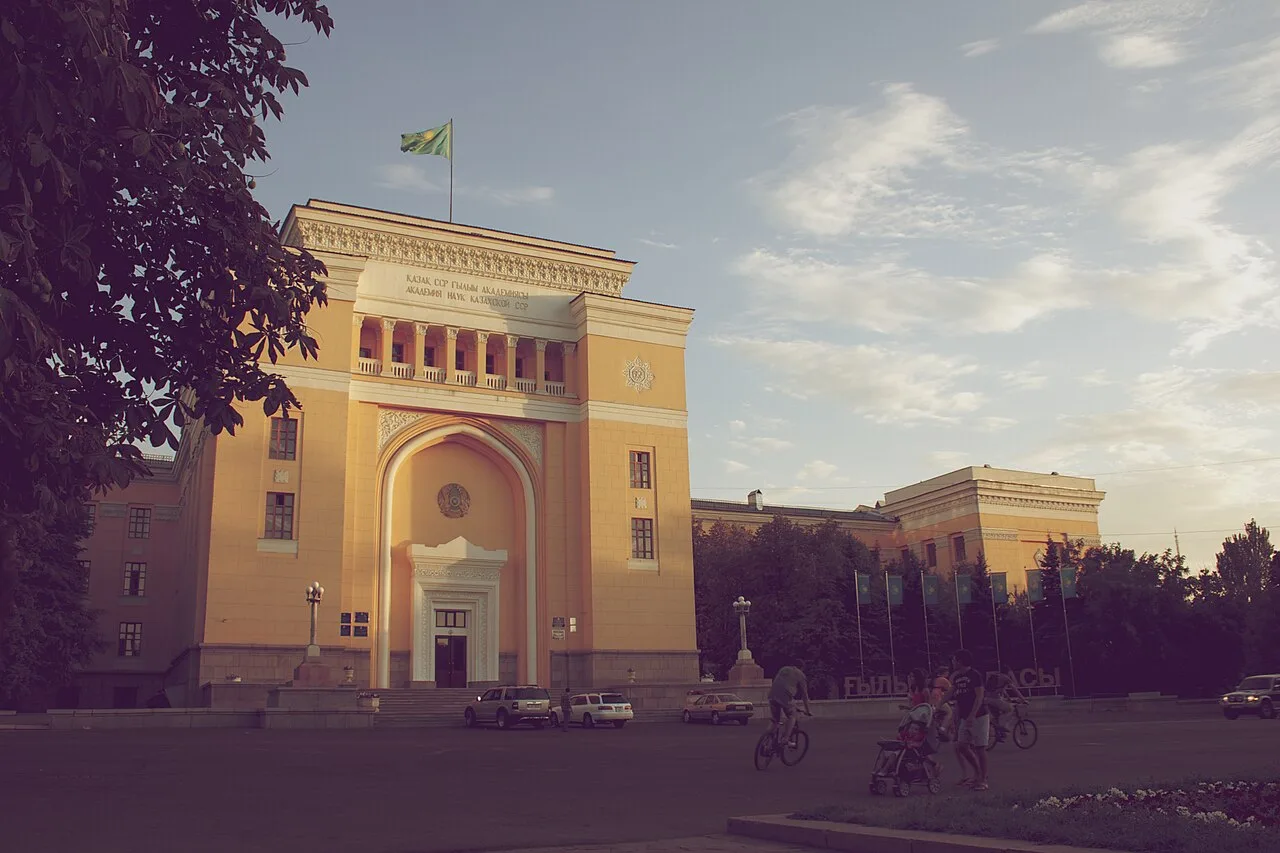 Академия наук на Шевченко, август 2012: жёлтый фасад в вечернем свете, высокая арка-пештак с белой дверью в глубине, над ней балкон с колоннами и надпись на двух языках. Справа уходят павильоны, перед ними флагштоки. Встаньте у клумбы на краю сквера — арка поместится в кадр целиком. Фото: Togzhan Ibrayeva, 2012, Wikimedia Commons, CC BY-SA 4.0