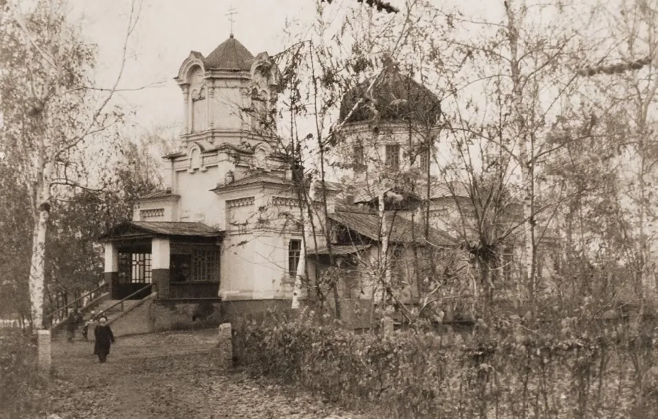 Казанский храм в 1944–1945 годах, архивное фото: деревянная церковь с куполами за голыми деревьями, крыльцо, у ограды одинокая фигура. Храм только что вернули верующим после десяти лет под замком. Фото: Митрополичий округ в Республике Казахстан, из книги «Казанский собор города Алматы», Wikimedia Commons, CC BY 4.0