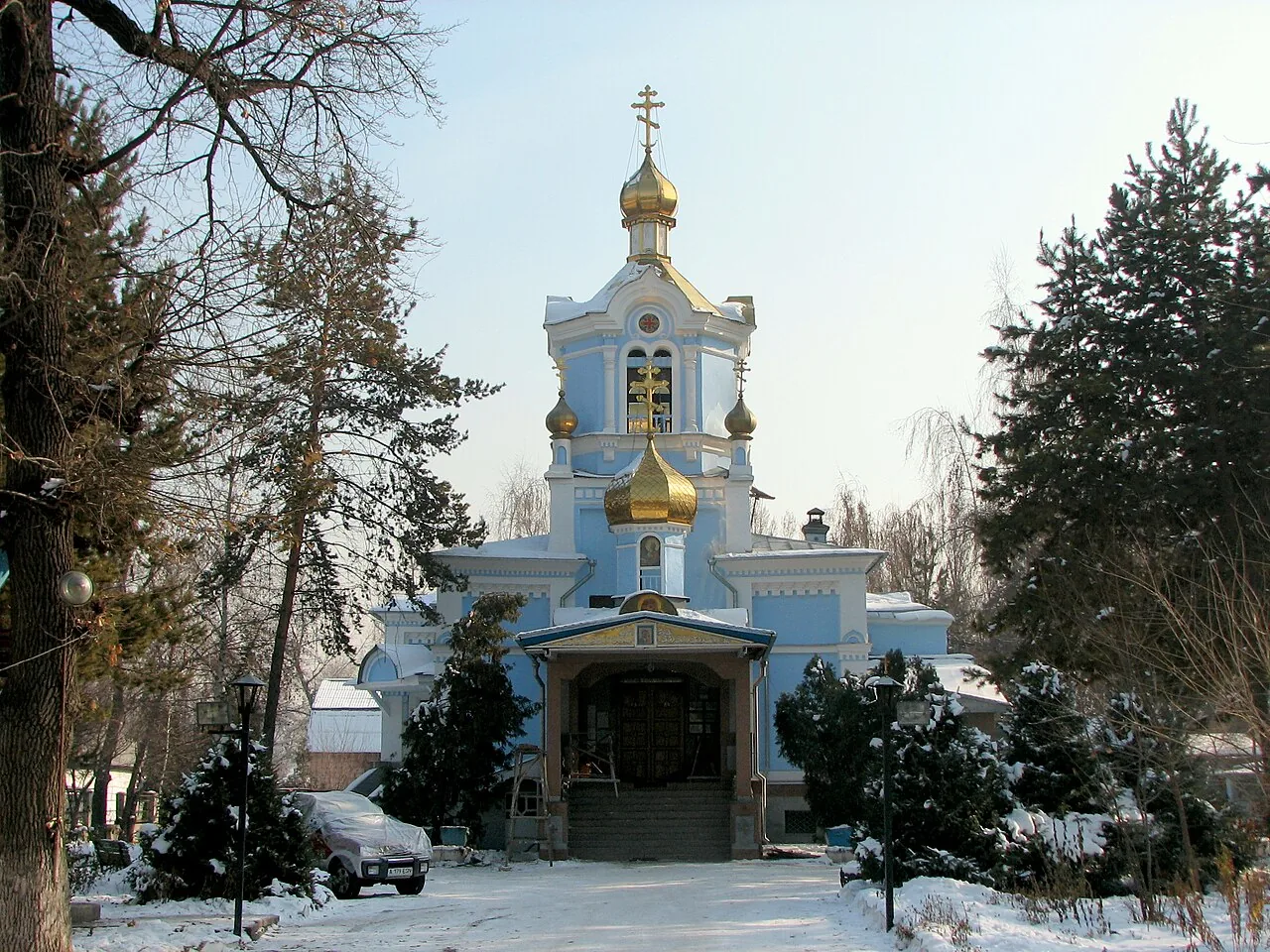 Казанский собор в декабре 2005 года: голубые стены, золотые купола с крестами, снег вдоль дорожки, ели и голые деревья вокруг, у ограды припаркованы машины. Зимой деревянный храм выглядит особенно лёгким, будто его можно поднять. Фото: Kefi, 2005, Wikimedia Commons, CC0