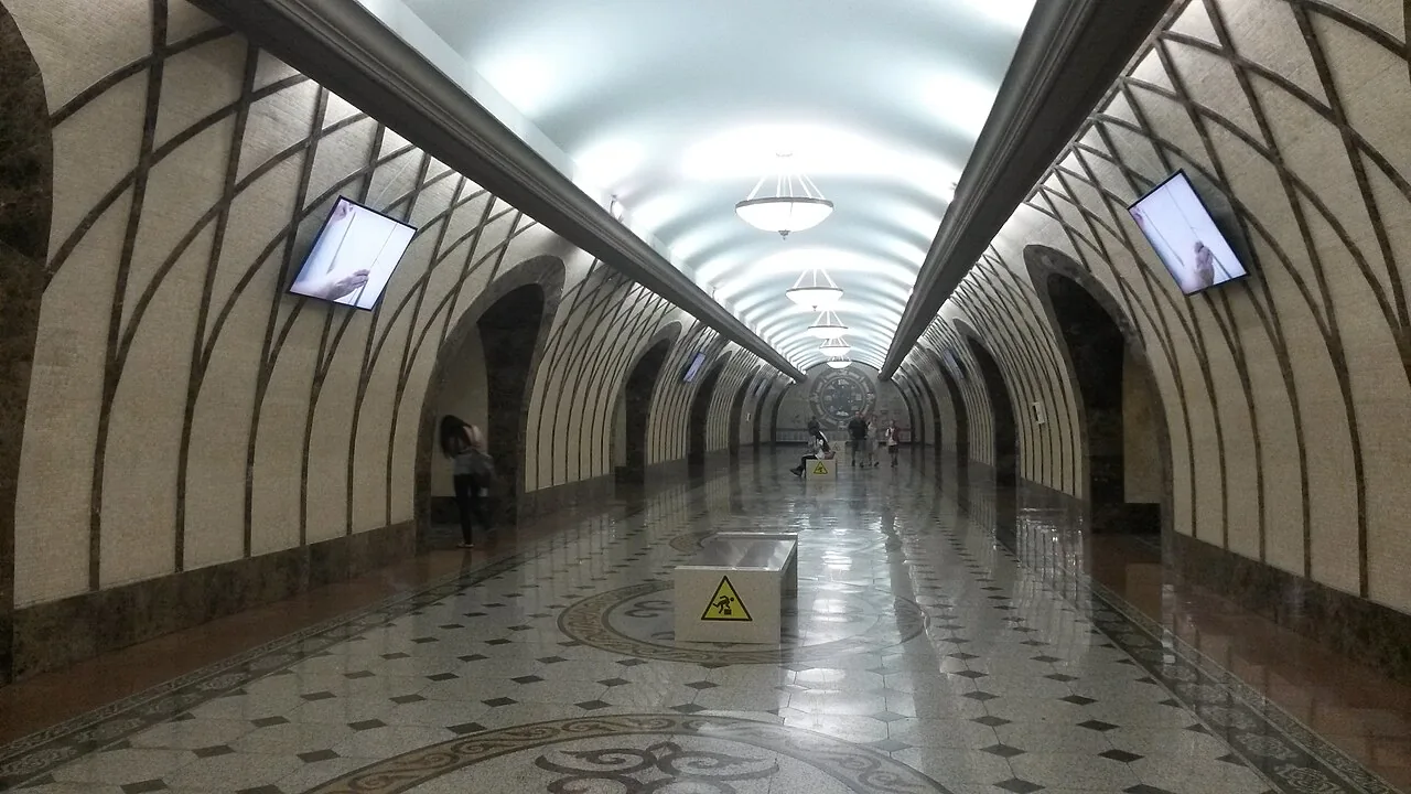 Платформа «Жибек Жолы», август 2014: своды в бежевой мраморной мозаике, тёмные дуги арок, гранитный пол с орнаментом, люстры уходят вдаль, как караван фонарей. С этого зала начинается прогулка. Фото: Alex J. Butler, 2014, Wikimedia Commons, CC BY 2.0