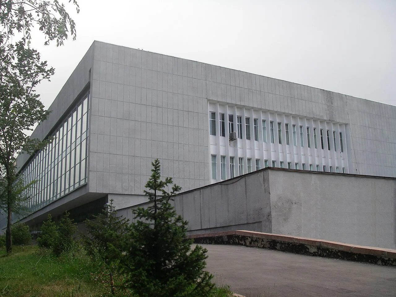 Угол Национальной библиотеки, июль 2010: белая плитка фасада, стеклянная лента второго этажа нависает над входом, справа — ритм узких окон, внизу каменный пандус и молодая ель. Встаньте у ели: угол здания поплывёт над пандусом, как корабль у пристани. Фото: Kimsemir, 2010, Wikimedia Commons, CC BY-SA 4.0