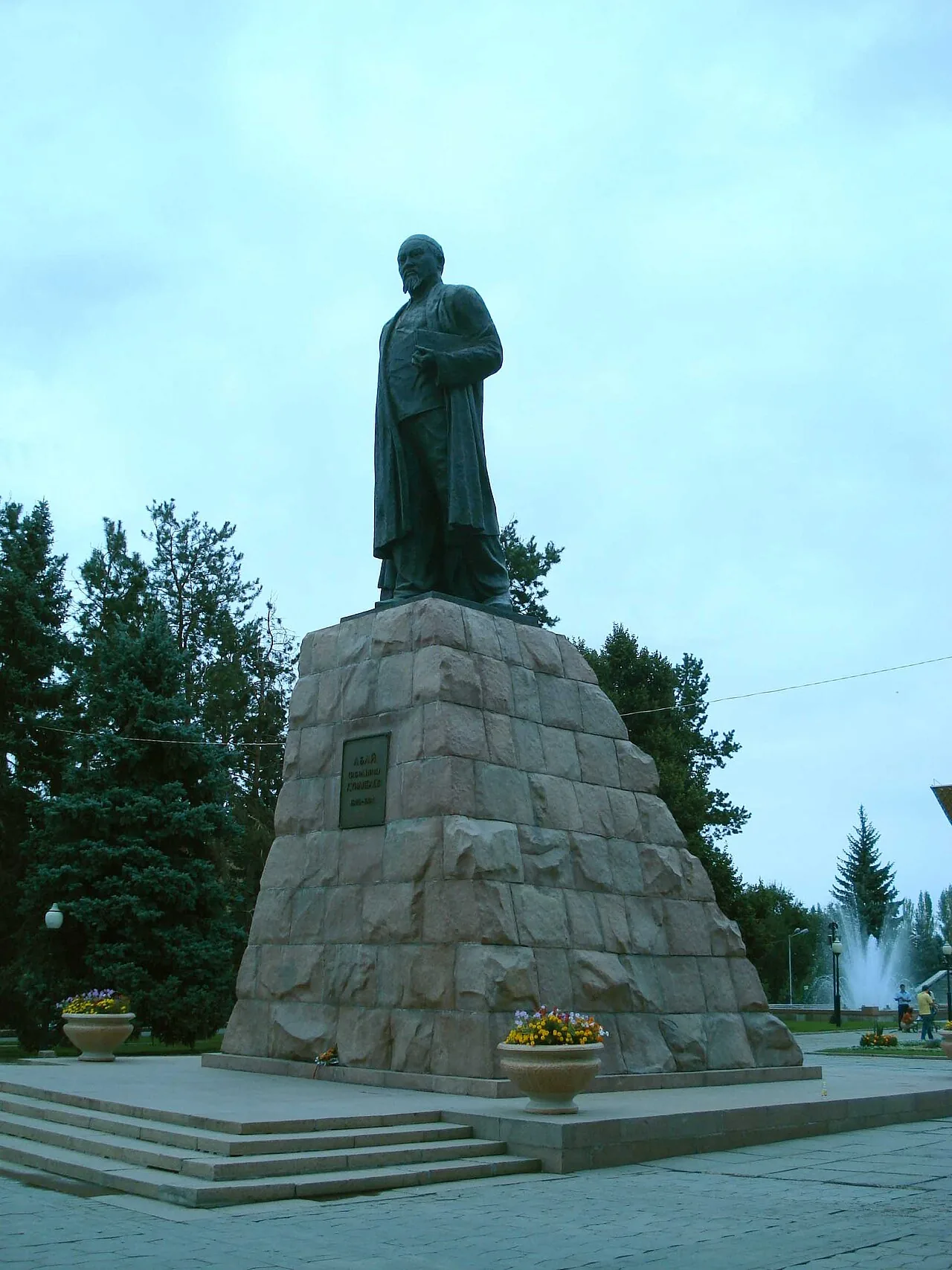 Памятник Абаю в мае 2008 года: бронзовая фигура на высоком постаменте из красного гранита, вокруг ели, у подножия вазоны с цветами, справа виден фонтан. Поищите в руке Абая книгу и сравните фигуру с постаментом: гладкий металл над рваным камнем. Фото: Mheidegger, Wikimedia Commons, общественное достояние