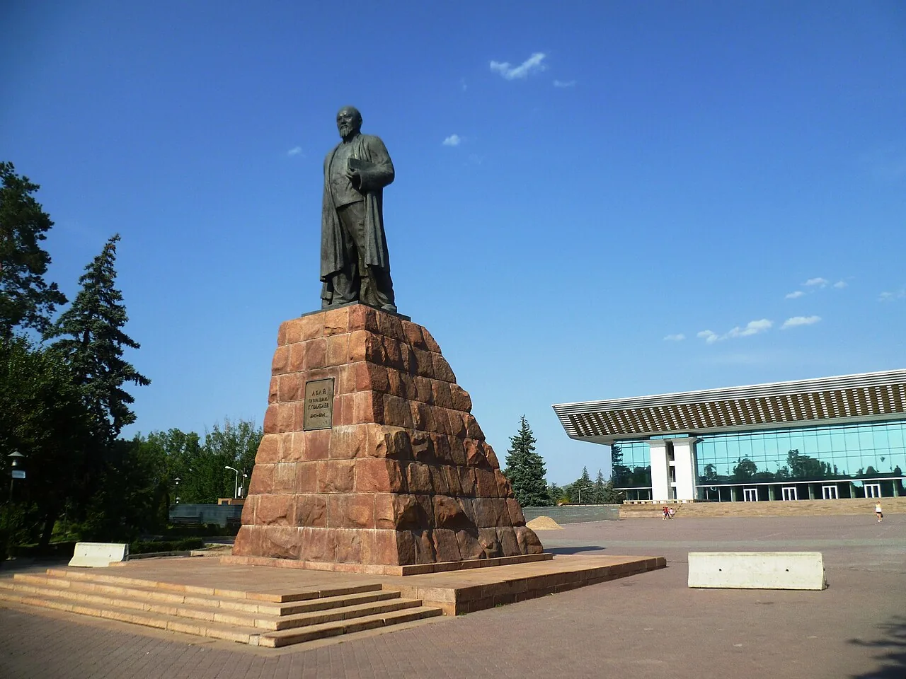 Памятник Абаю на площади Абая, август 2017: тёмная бронзовая фигура на ступенчатом постаменте из красного гранита, за ним длинное здание со стеклянным фасадом, синее небо и ели. Памятник открыли в 1960 году; высота — тринадцать метров. Фото: Radosław Botev, Wikimedia Commons, CC BY 3.0 pl