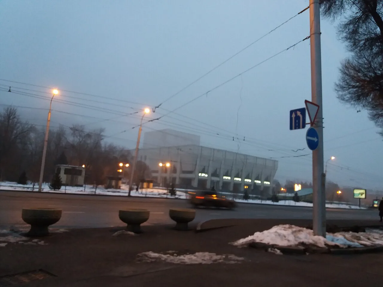 Театр имени Ауэзова в феврале 2019 года, в сумерках: здание за деревьями, фонари, провода над проспектом, остатки снега у бордюра. Так он выглядит в конце нашей прогулки, если идти зимой. Фото: Мазур Владимир, Wikimedia Commons, CC BY-SA 4.0