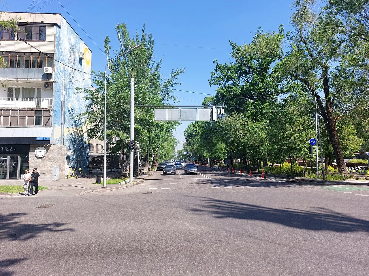 Улица Кунаева от Курмангазы, апрель 2026: широкая улица в зелени, на торце дома слева — огромный мурал в синих завитках, как «Звёздная ночь», ниже витрина и часы. Центр, по которому идём к малой сцене, давно рисует на своих стенах. Фото: Almatrkh, 2026, Wikimedia Commons, CC BY 4.0