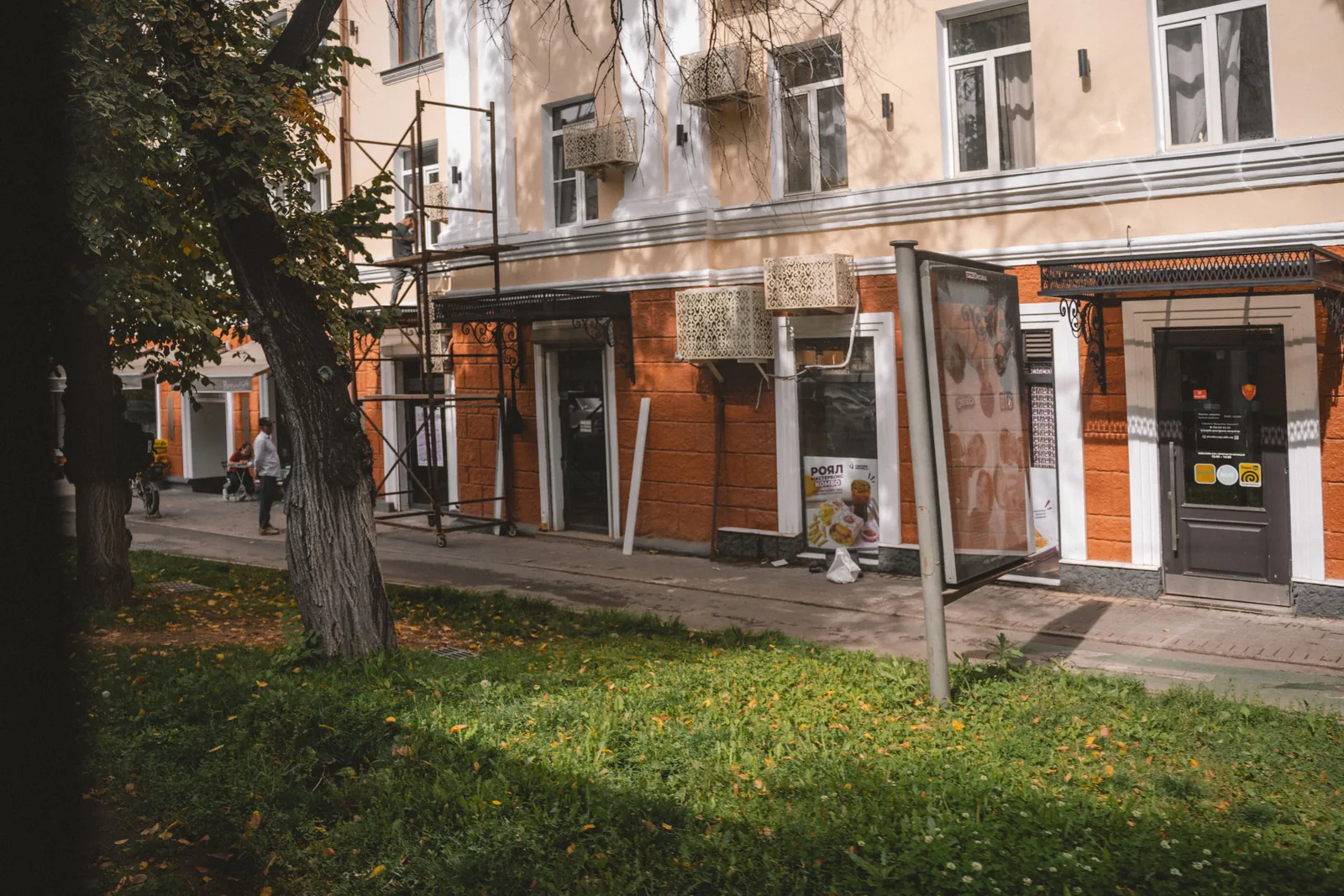 Фасад старого дома в центре, осень: двухэтажное здание со светлыми стенами и высокими окнами, перед ним газон и молодое дерево, на углу дверь подъезда. Типичный дом квартала, за которым прячется двор. Фото: Валентин Шафранов