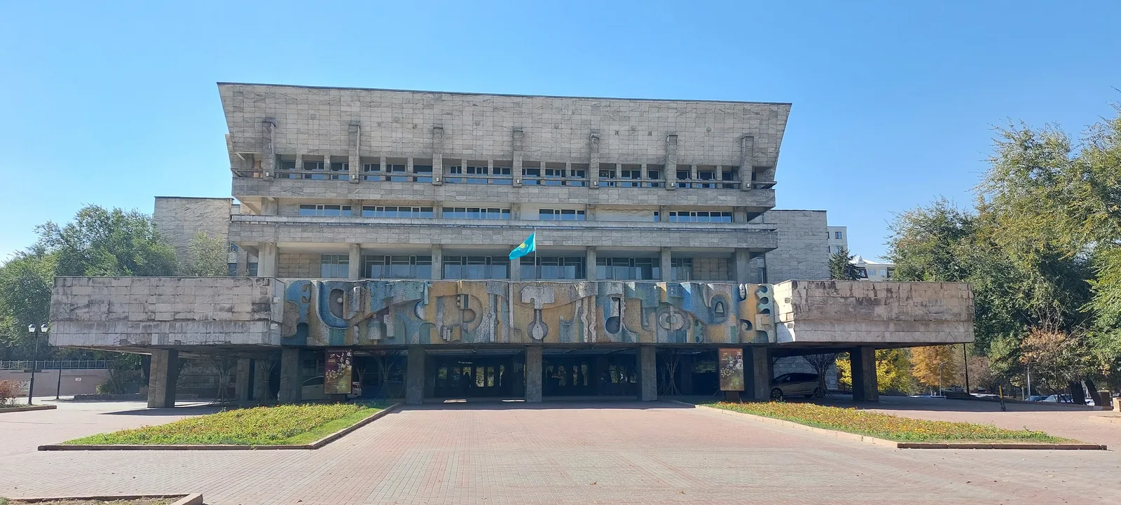 Здание советского модернизма в центре Алматы, солнечный осенний день: над входом тянется рельефный фриз — трубы, круги, клавиши, всё в синем, охре и сером. Встаньте у клумбы и найдите, где рельеф повторяется, а где художник нарушил ритм. Фото: Валентин Шафранов