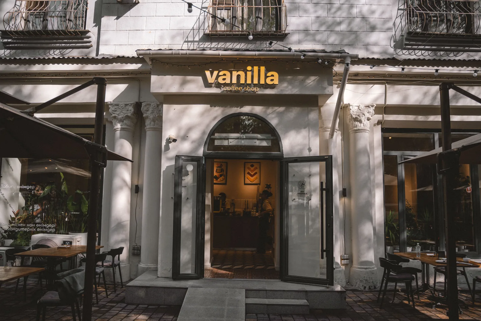 Вход в одну из кофеен Vanilla: белый фасад старого дома, колонны с резными капителями, арка над дверью, гирлянда лампочек и тень листвы на стене. Двери открыты — кофейня приглашает раньше, чем вы решили зайти. Фото: Валентин Шафранов