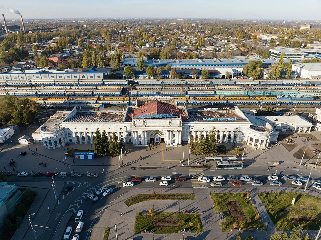Вокзал Алматы-2 сверху, ноябрь 2022: белое здание с колоннадой и крыльями, за ним ряды вагонов на путях, перед ним площадь с газонами и парковкой, вдали — крыши и деревья. Отсюда видно, как вокзал стоит на краю старого города, лицом к проспекту. Фото: Nikolai Bulykin, Wikimedia Commons, CC BY-SA 4.0