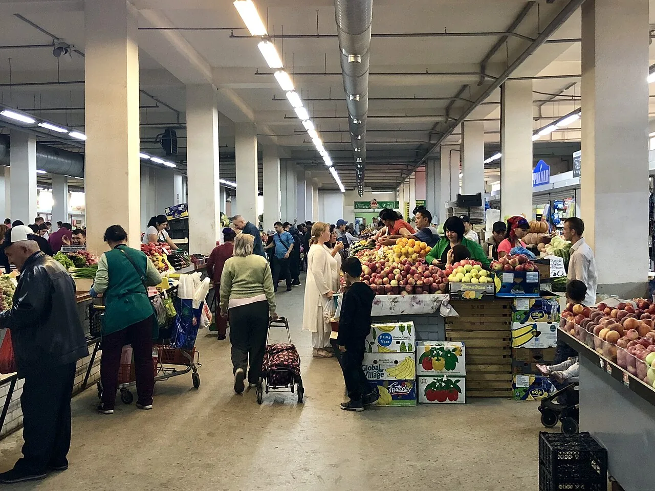 Проход между рядами главного зала, сентябрь 2019: колонны, лампы под потолком, ящики с фруктами, покупатели с тележкой. Под этой крышей дождь слышен только как далёкий стук сверху. Фото: Davide Mauro, 2019, Wikimedia Commons, CC BY-SA 4.0